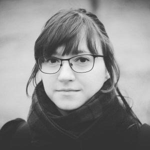 Schwarz-Weiß Fotografie von Mika M. Krüger. Sie trägt eine Brille, Schal und einen Zopf. 